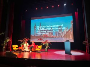 Photo shows brightly it stage with The 13th International Club Health Conference screen in the background. Two women are discussing things on the stage.